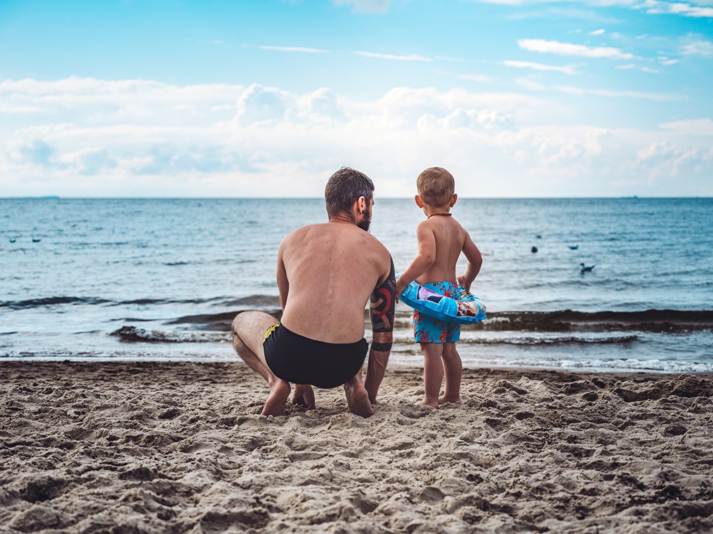 We see a father and son on the beach looking out at the sea in the image.
